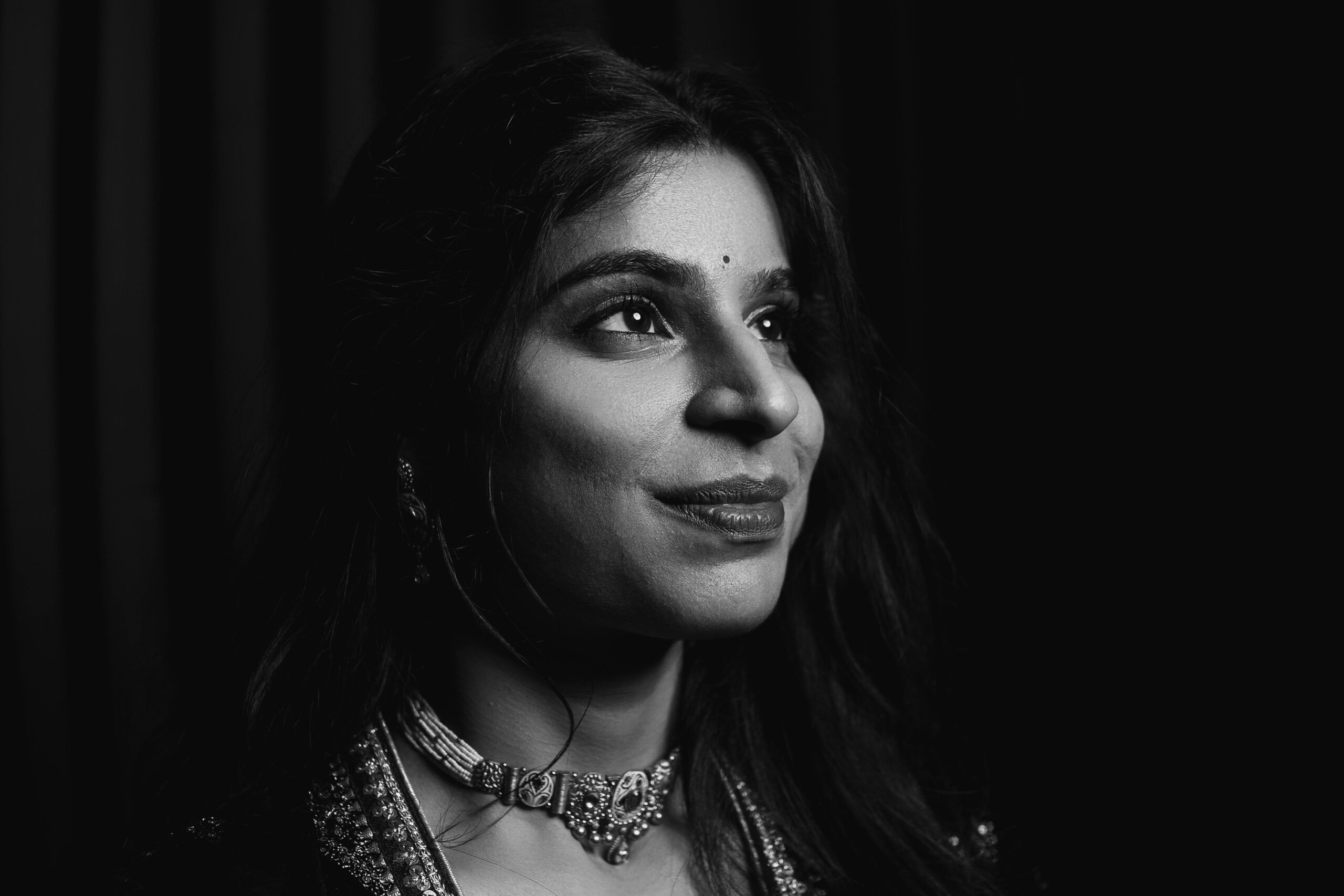 Captivating black and white portrait of a woman with a necklace against a dark background.