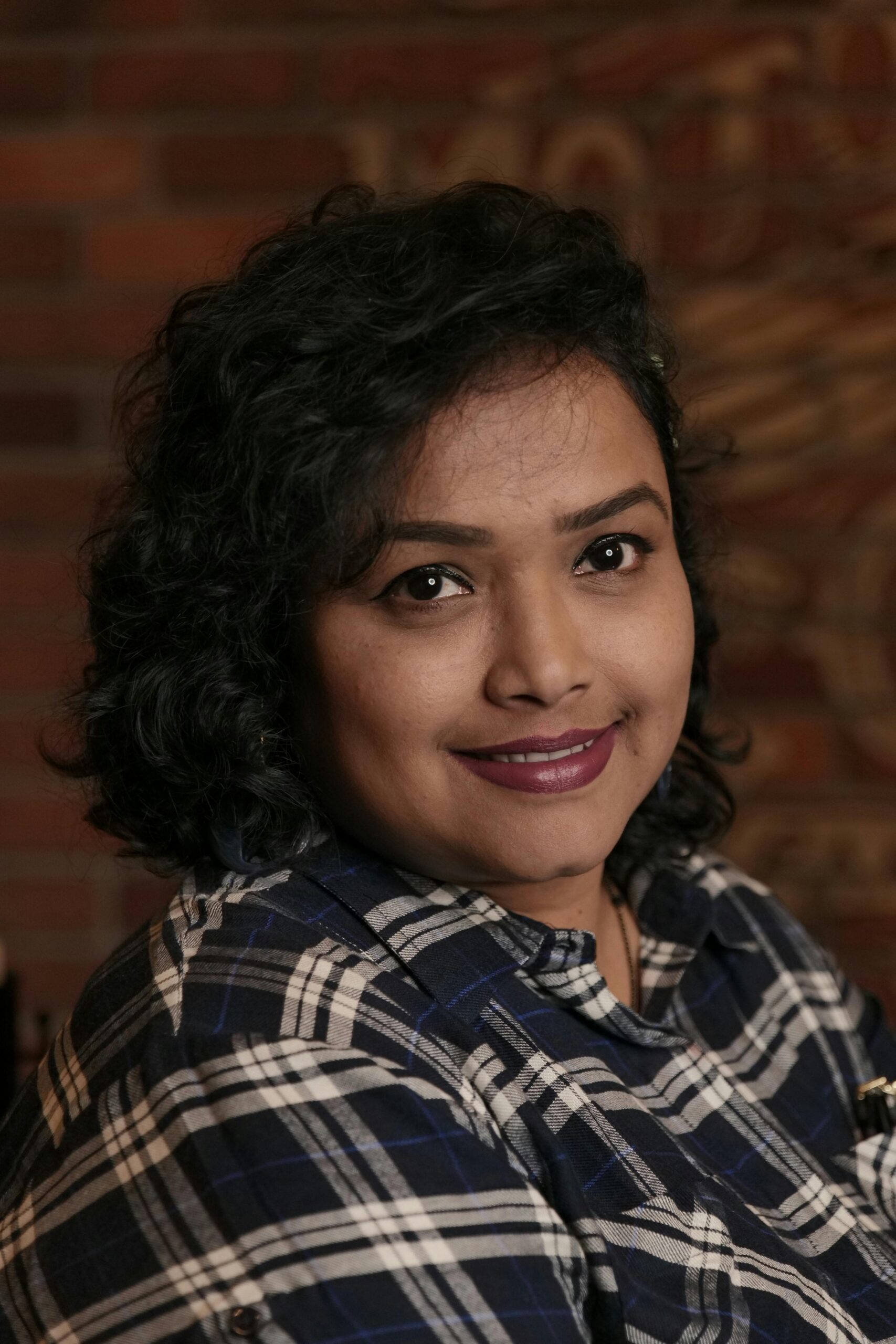 A portrait of a smiling woman with curly hair in a plaid shirt against a brick wall background.